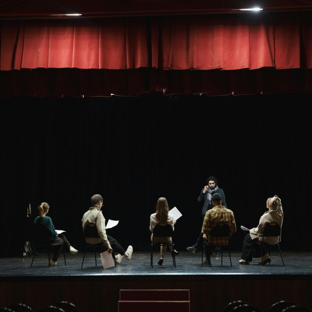 Répétition théâtrale sur scène, un metteur en scène face à des comédiens assis en demi-cercle devant le rideau rouge